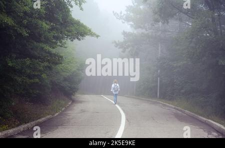 Straße in nebligen Wald, einsame Mädchen Wanderer geht weg in unheimlichen Wäldern, verloren junge Frau Reisende ist allein im Nebel. Landschaft von gruseligen Ort. Konzept von h Stockfoto