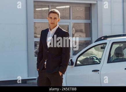 Junger männlicher Berater in Autoschau, der in der Nähe von Autos steht und die Kamera anschaut Stockfoto