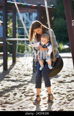 Verbringen Sie ein wenig Zeit im Freien. Junge Mutter auf einer Parkschaukel mit einem kleinen Jungen. Stockfoto