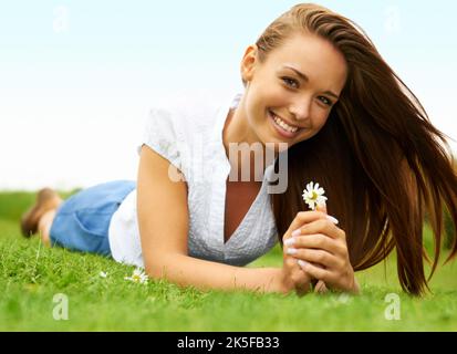 Frisch gesichtige junge Schönheit. Nette junge Frau lächelt und hält eine Blume, während sie auf dem Gras liegt. Stockfoto
