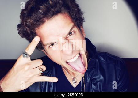 Rock'n Roll. Nahaufnahme eines jungen Mannes, der seine Zunge herausstreckt und Handgesten macht, umgeben von einem Lichteinfall. Stockfoto