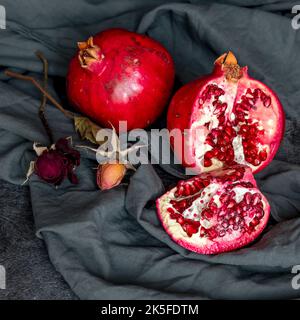 Zwei rote Granatäpfel, ein ganzer und der andere, in zwei Hälften geschnitten, daneben zwei Rosen, eingewickelt in einen grauen Stoff Stockfoto