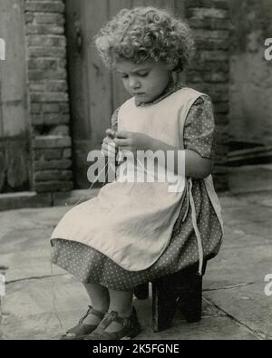 Ein kleines Mädchen mit lockigem Haar trägt ein gepunktetes Kleid, Schürzenkleid und Sandalen, während sie in einem mit Steinplatten gepflasterten Hof strickt sitzt. Vintage-monochrome Porträtfotografie Ende 1930s von Kathleen Blenkhorn ARPS, einer Fotografin aus Birkby, Huddersfield, West Yorkshire. Das Bild wurde mit einer Rolleiflex TLR-Kamera (Twin Lens Reflex) mit einem Tessar F3,5 ‘Taking’ Objektiv auf mittelformatem Schwarz-Weiß-Film aufgenommen. Stockfoto