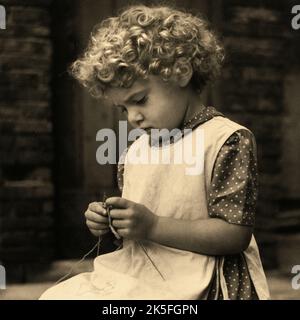 Ein junges Mädchen mit einem Mopp von Locken trägt ein gepunktetes Kleid und Schürze, während sie strickt sitzt. Sepiafarbene quadratische Details einer monochromen Vintage-Porträtfotografie von Kathleen Blenkhorn ARPS aus Birkby, Huddersfield, West Yorkshire. Das Originalbild wurde mit einer Rolleiflex TLR-Kamera (Twin Lens Reflex) mit einem Tessar F3,5 ‘Taking’-Objektiv auf mittelformatem Schwarz-Weiß-Film aufgenommen. Stockfoto