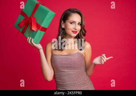 Charmante, lächelnde elegante Frau im Galadress halten Geschenk-Box, zeigen etwas mit Zeigefinger auf roten Studio-Hintergrund. Stockfoto