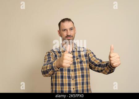 Attraktiver, fröhlicher, bärtiger hispanic-Mann mit kariertem Hemd, isoliert auf beigem Hintergrund stehend und Daumen nach oben zeigend. Stockfoto