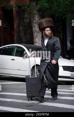 Ein orthodoxer jüdischer Mann, der sich auf den Sabbat vorbereitet, trägt einen Shtreimel-Pelzhut und überquert mit einem Koffer die Straße. In Williamsburg, Brooklyn, New York. Stockfoto