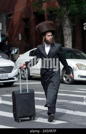 Ein orthodoxer jüdischer Mann, der sich auf den Sabbat vorbereitet, trägt einen Shtreimel-Pelzhut und überquert mit einem Koffer die Straße. In Williamsburg, Brooklyn, New York. Stockfoto