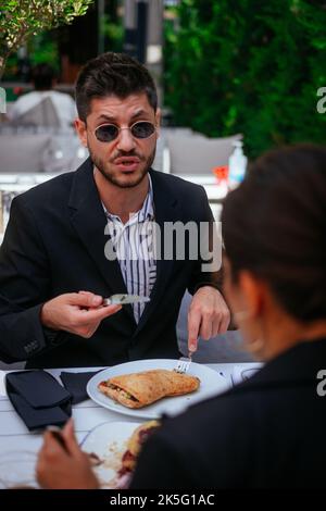 Ein gut aussehender, verärgerter Geschäftsmann frühstückt, während er wütend auf das Projekt ist und versucht, dies seiner Kollegin zu erklären Stockfoto