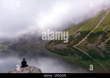 Friedliche Landschaft in der polnischen Tatra mit einem See und einem Mann, der auf einem großen grauen Bergfelsen sitzt Stockfoto