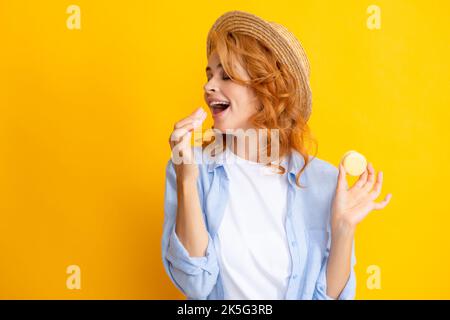 Nette junge Frau halten Makronen. Schönes lächelndes junges Mädchen, das bunte Makronen auf gelbem Hintergrund isst. Stockfoto