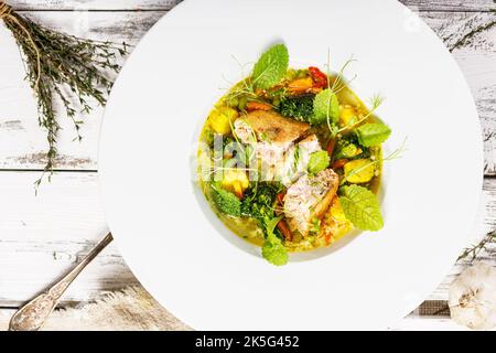 Draufsicht Lachsschnitzel mit Fischbrühe und Gemüse. Appetitliche Fischfleischbällchen mit Gemüsesuppe. Serviert auf einem weißen Teller. Weißer Hintergrund aus Holz Stockfoto