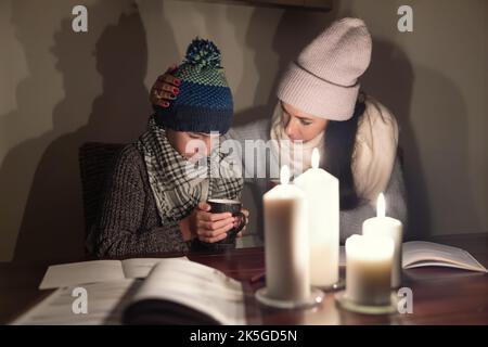 Besorgte Mutter tröstet ihren eiskalten Sohn mit Tee und studiert bei Kerzenlicht in der Kriegszone, beide tragen Winterkleidung zu Hause. Stockfoto
