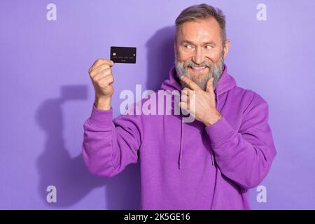 Portrait von alten positiven guten Laune fröhlichen Mann gekleidet lila Hoodie Blick auf schwarze Kredit-Debit-Karte isoliert auf violetten Hintergrund Stockfoto