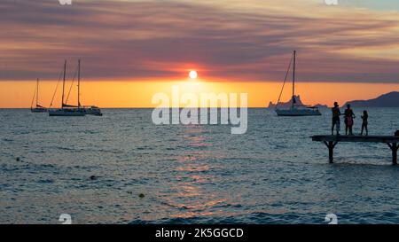 Drei Freunde am Pier beobachten den Sonnenuntergang über dem Meer und Segelboote vor Anker auf der Insel Formentera in Spanien