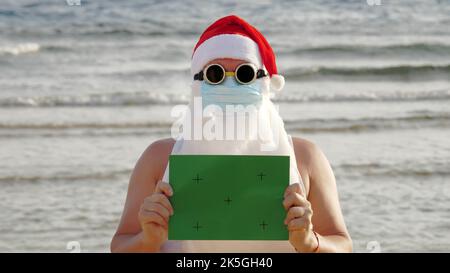 Witziger weihnachtsmann, in einer Schutzmaske. Santa, in Sonnenbrille, mit einem grünen Schild, vor dem Hintergrund des Meeres. santa claus Urlaub während Coronavirus Pandemie oder Lockdown, an der Küste. Hochwertige Fotos Stockfoto