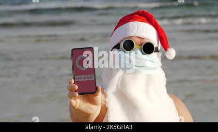 Witziger weihnachtsmann, in einer Schutzmaske. Santa, in Sonnenbrille, hält Smartphone mit COVID-19-Symbol, Text - Pandemie, auf dem Bildschirm, am Strand am Meer. santa claus warnt vor Coronavirus Gefahr. Hochwertige Fotos Stockfoto