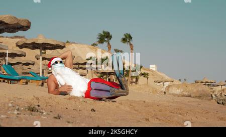 Weihnachtsmann Sommerurlaub. Weihnachtsmann mit Spaß. Lustiger Weihnachtsmann, in Schutzmaske, Sonnenbrille und Flossen, entspannt am Sandstrand am Meer, unter Palmen. Hochwertige Fotos Stockfoto