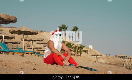 Weihnachtsmann Sommerurlaub. Weihnachtsmann mit Spaß. Lustiger Weihnachtsmann, in Schutzmaske, Sonnenbrille und Flossen, entspannt am Sandstrand am Meer, unter Palmen. Hochwertige Fotos Stockfoto