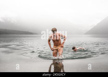 Sportlicher kaukasischer Junge ohne Kleidung und mit einem Badeanzug, der nass und müde aus dem Wasser kommt und mit seinem Freund im kalten Wasser schwimmt Stockfoto
