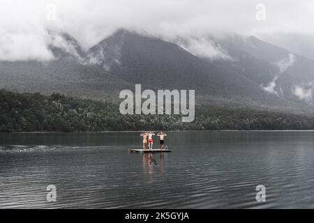 Drei kaukasische Jungen stehen in Badehosen gekleidet und scherzen von der hölzernen Plattform in der Mitte des Sees im ruhigen Wasser mitten in Stockfoto