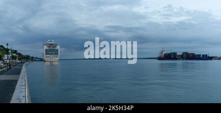 Cobh, Irland - 15. August 2022: Panoramablick auf ein großes Kreuzschiff auf dem Pier in Cobh mit einem großen Frachtschiff, das in der Dämmerung in den Hafen von Cork einfährt Stockfoto