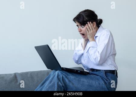 Stilvolle schockierte junge Frau mit Laptop in den Händen auf dem Sofa und Blick auf Laptop. In einem weißen Hemd und Jeans gekleidet. Graue Wand mit Schatten hinter ihr. Stockfoto