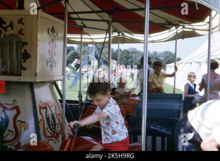 East Sussex. 1964. Kleine Kinder reiten auf einer fröhlichen Runde auf einem Landmarkt. Das Karussell ist handbemalt und mit Miniatur-Motorwagen und Motorrädern ausgestattet. In der Ferne sind eine Schaukelbootfahrt und ein großes Festzelt zu sehen. Stockfoto