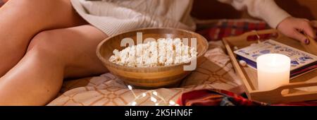 Gesichtslose Frau in Strickwolle kuscheliger Pullover im Bett auf einer Decke im Camping-Wohnwagen mit einem Buch und Popcorn-Essen und Genuss auf glücklichem Neuem Stockfoto