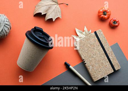 Kaffeebecher mit Naturkorkband. Kaffeebecher zu trockenen Platanenblättern und Deko-Kürbissen. Notizbuch mit passendem Korkbezug. Flach auf orange legen Stockfoto