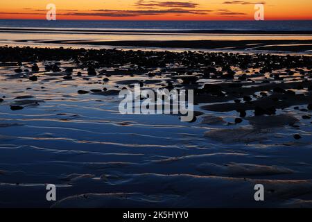 Der Sonnenaufgangshimmel spiegelt sich im seichten Wasser und dem welligen und felsigen Sand bei Ebbe am Strand von Cape Cod wider Stockfoto