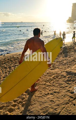 Ein Mann mittleren Alters trägt sein Surfbrett an einem sonnigen Sommerferientag am Waikiki Beach in Hawaii zum Ufer und den Wellen Stockfoto