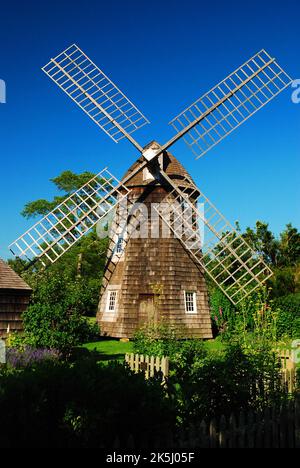 Eine historische hölzerne Windmühle befindet sich inmitten eines blühenden Sommergartens in einem Museum, das der Darstellung des ländlichen Koloniallebens gewidmet ist Stockfoto