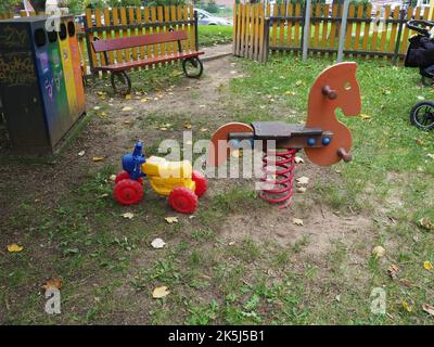 Buntes Plastikmotorrad und ein Schaukelspielzeug aus Holz auf einem Spielplatz mit Holzbank, Holzzaun und Abfalleimer im Hintergrund Stockfoto