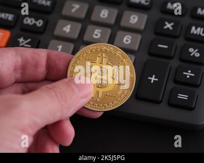 Ein Mann mit einer goldenen Bitcoin-Münze in der Hand und einem Rechner im Hintergrund. Nahaufnahme. Stockfoto