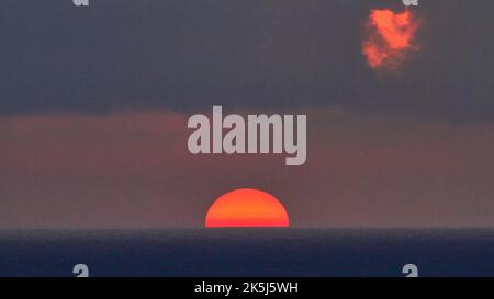 Sonne als feuerrote Großkugel, blau-violetter Himmel, einzelne Wolke in Rot beleuchtet, Sonne direkt hinter dem Horizont, halb versunken, Sonnenuntergang, Abendlicht Stockfoto