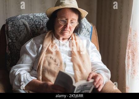 Ältere Frau, die ein Buch liest, sitzt auf dem Stuhl am Fenster Stockfoto