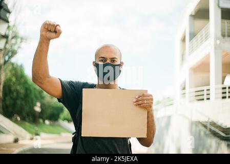 Ein dunkelhäutiger junger Mann in einer schwarzen Maske hält ein leeres Plakat in der einen Hand, während die andere Hand zu einer Faust über seinem Kopf zusammengeballt ist. Stockfoto