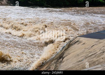 Schlammige Wasserströme, die nach einem Regensturm schnell auf einen Fluss rauschen. Die Stromschnellen, die über ein Wehr hinunterfallen, spritzen, sprühen, sprühen und blasen. Stockfoto