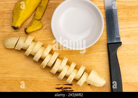 Draufsicht, flache Scheibe roher reifer, organisch geschälter Banane mit Küchenmesser und weißem Keramikbecher auf Holzschneidebrett. Stockfoto