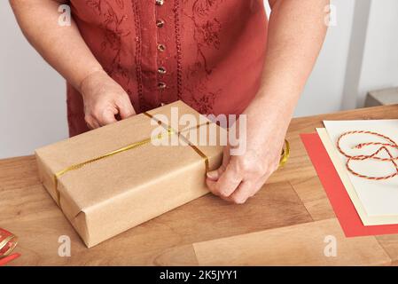 Unkenntlich reife Frau, die ein Weihnachtsgeschenk mit neutralem Papier und einem goldenen Zierband verpackt. Stockfoto