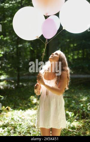 Sich kindlich fühlen. Eine hübsche junge Frau mit einem Haufen Ballons im Wald. Stockfoto