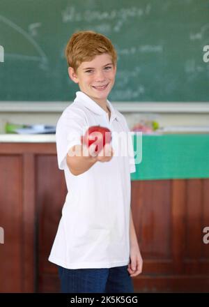 Hier ist ein Apfel für Sie. Netter kleiner Schuljunge, der einen Apfel im Klassenzimmer hält. Stockfoto