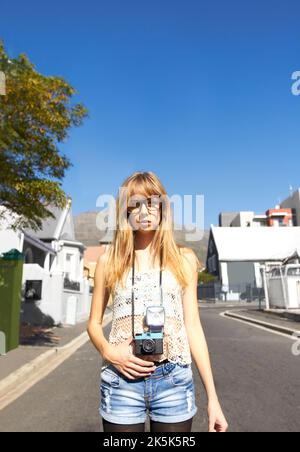 Malerischer, urbaner Stil. Ein stilvolles junges Mädchen mit einer Vintage-Kamera, das mitten auf einer Straße in einem malerischen Wohnviertel steht. Stockfoto