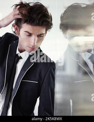 Er weiß zu posieren. Ein junger Mann, der sich mit der Hand im Haar gegen die Klasse lehnt. Stockfoto