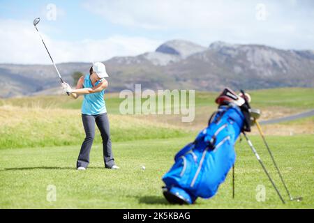 Den Schnitt machen. Eine Golfspielerin, die gerade einen Schuss mit einer verschwommenen Golftasche im Vordergrund machen wird. Stockfoto