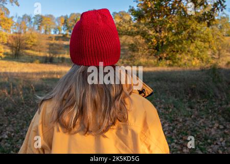 Junge Frau mit langen blonden Haaren in einem roten Hut und gelbem Mantel, die im Herbstpark spazieren geht. Rückansicht. Leuchtende Herbstfarben. Sonniges Wetter. Genießen Sie das Leben Stockfoto