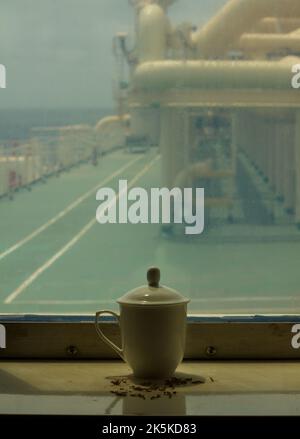 Blick auf eine weiße Teetasse mit Deckel in der Nähe des Fensters eines LNG-Schiffes auf See Stockfoto