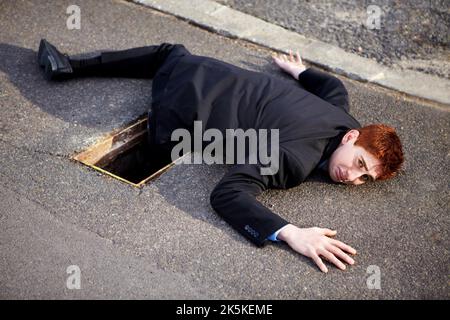Einige Unternehmungen bringen Sie in ein Loch. Ein unglücklicher Geschäftsmann, der auf dem Boden lag und sein Bein in einem Abfluss steckte. Stockfoto