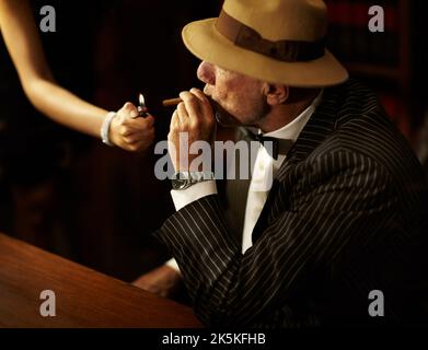 HES bekam Macht und Einfluss über andere. Der gealterte Mafioboss trägt einen Hut und sieht ernst aus, während eine Frau eine Zigarette für ihn anzündet. Stockfoto